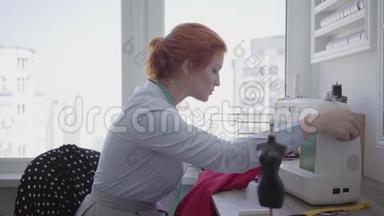 专业漂亮的红发女人在车间的桌子上缝衣服。 裁缝工作勤奋，细心. 业余爱好